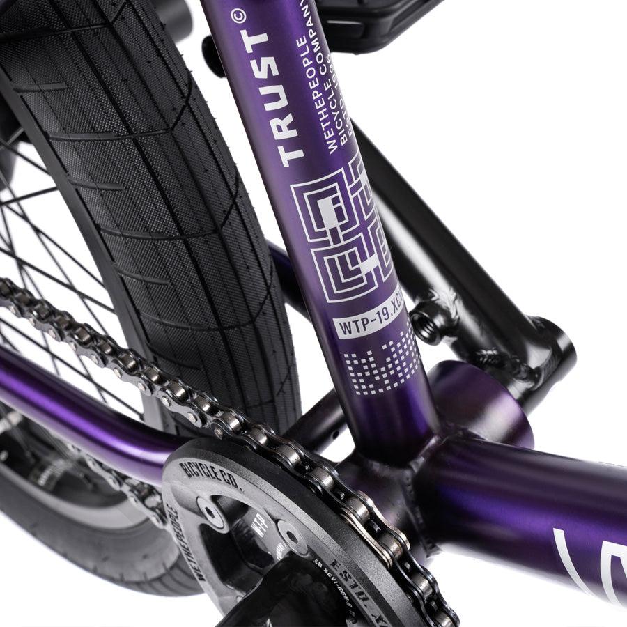 Close-up of a Wethepeople Trust 20 Inch Cassette/Hybrid Bike’s purple frame, showing the chain, rear tire, sprocket, "TRUST" branding, and Eclat Shift Freecoaster Hub alongside top-tier technical features.