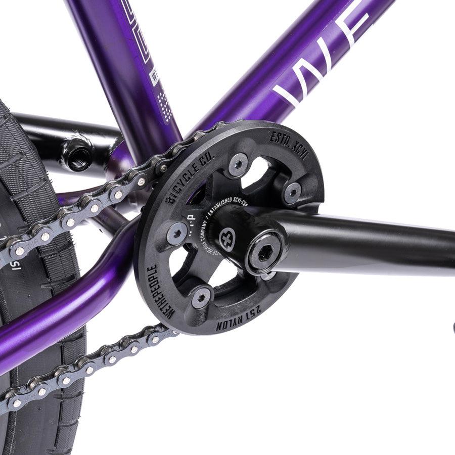 Close-up view of the Wethepeople Trust 20 Inch Cassette/Hybrid Bike drivetrain, featuring a black sprocket, chain, crank arm, and part of the purple frame.