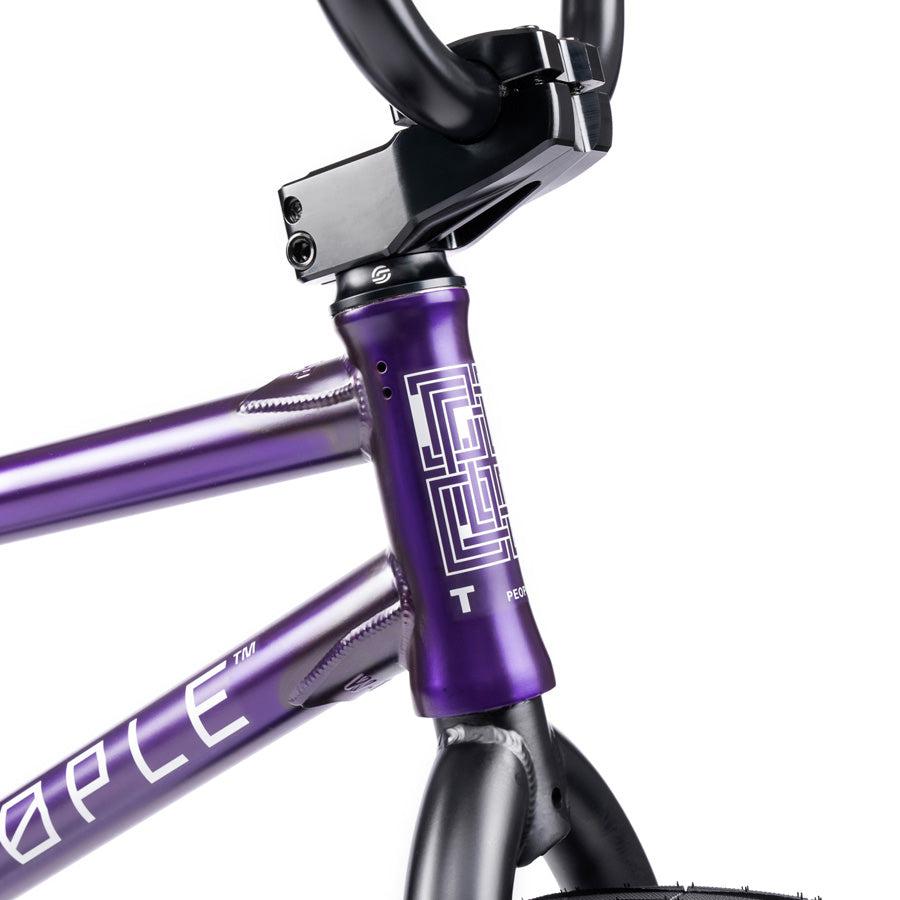 Close-up of a purple Wethepeople Trust 20 Inch Cassette/Hybrid Bike frame, highlighting the head tube, top tube, and handlebars with a geometric design on the head tube.