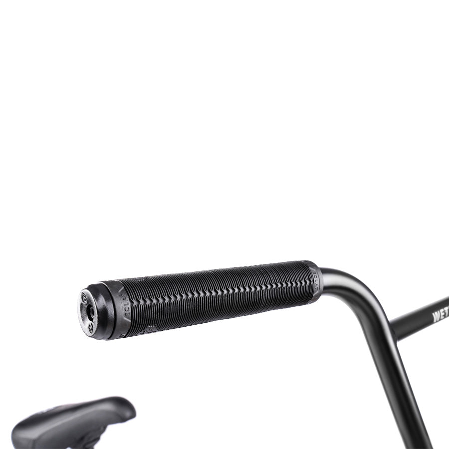 Close-up of a black textured handlebar grip on the Wethepeople Trust 20 Inch Cassette/Hybrid Bike, attached to a black handlebar; part of the bicycle seat is visible in the lower left corner.