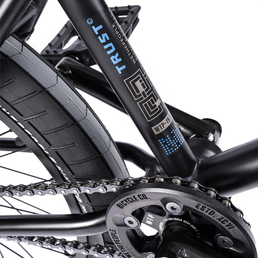 Close-up of the frame from the Wethepeople Trust 20 Inch Freecoaster/Hybrid Bike in black, showcasing the Trust brand logo, chain, and part of the rear wheel with its unique battleship geometry.