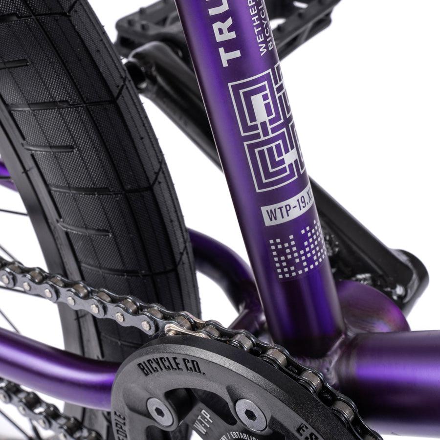 Close-up of the rear frame of the Wethepeople Trust 20 Inch Freecoaster/Hybrid Bike, showcasing its chain, tire, and branded decal on the purple frame.