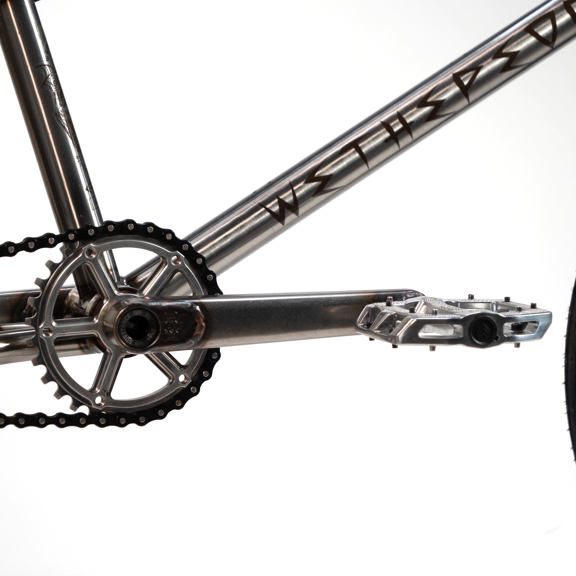 Close-up view of a bicycle crankset and pedal attached to a metallic bike frame with visible chainring and chain, part of the sleek design of the Wethepeople X LUX LAB 'Ironhide' Chaos Machine 20 Inch Custom Bike.