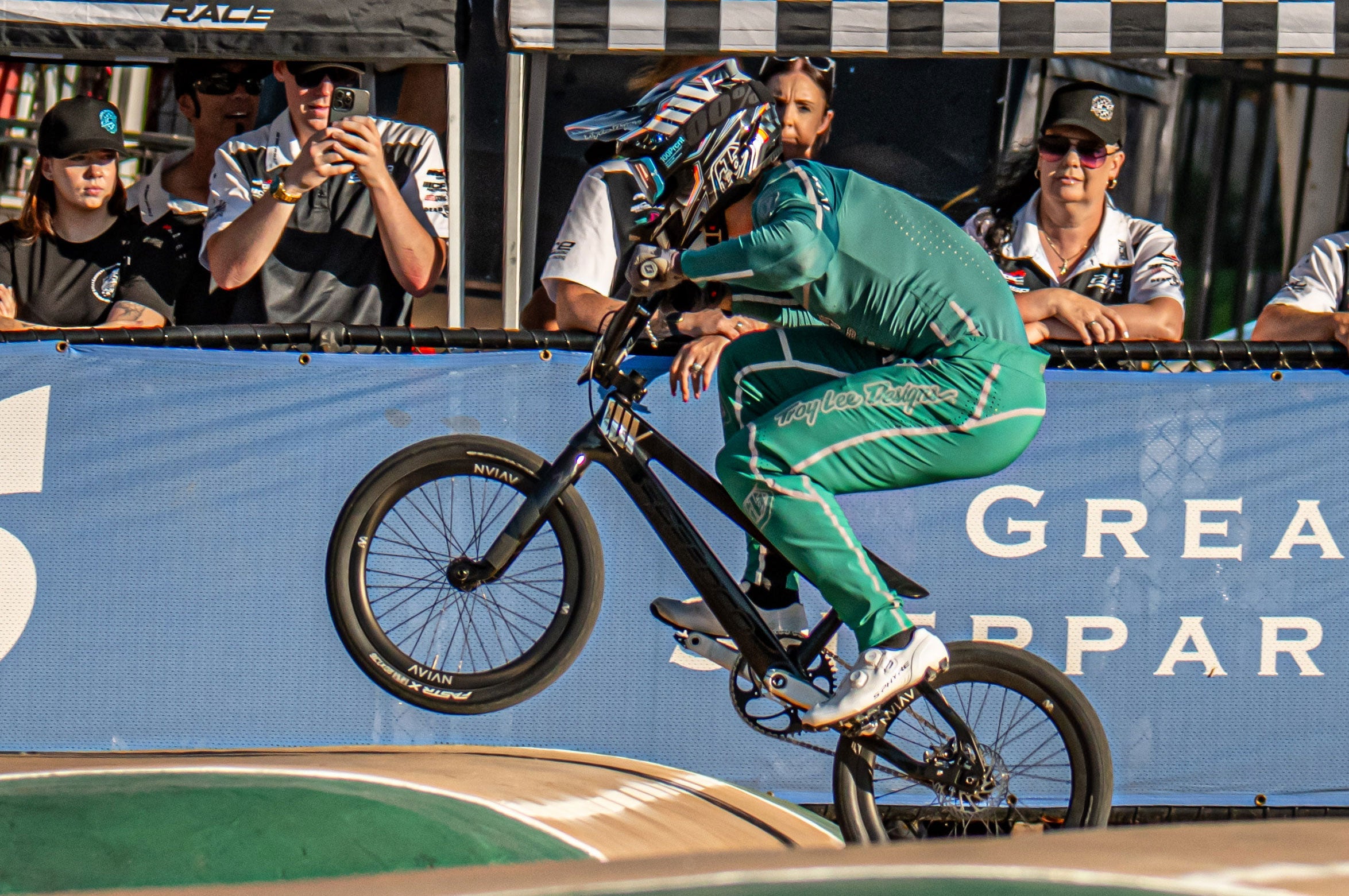 A cyclist in a green suit and helmet rides a BMX bike over a ramp during a race, with spectators watching in the background.