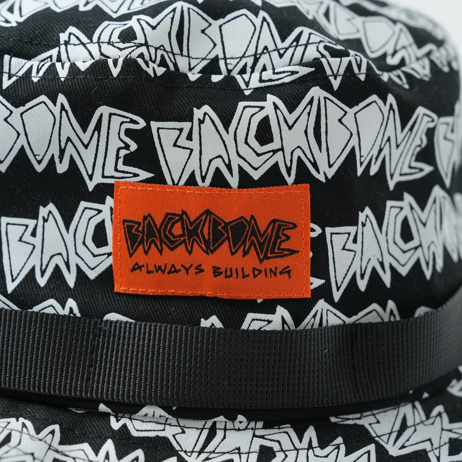 Close-up of a Backbone AO Boonie, showcasing a white "BACKBONE" graphic pattern and an orange patch with the words "BACKBONE ALWAYS BUILDING.