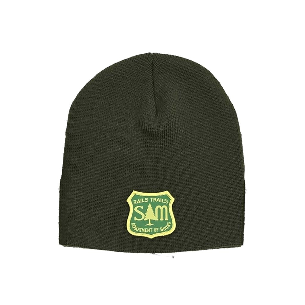 The S&M Department Of Freestyle Beanie is a dark green, knit beanie adorned with a biking patch, featuring a yellow tag that has "S&M Rails Trails Department of Birds" text and a tree silhouette, and is crafted from hypoallergenic acrylic.