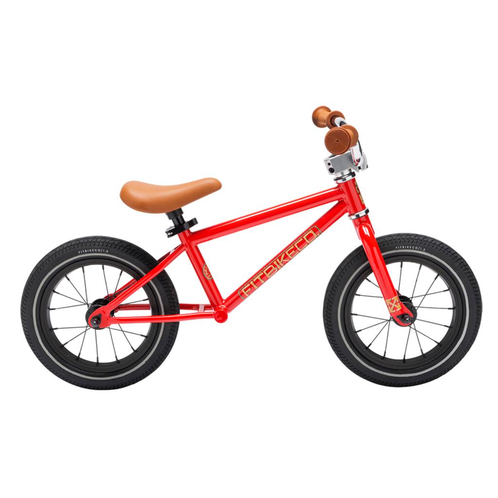The Fit Misfit 12 Inch Balance Bike is a small red BMX balance bike with a sturdy TIG welded steel frame, black tires, and tan seat and handlebar grips, shown in profile on a white background.