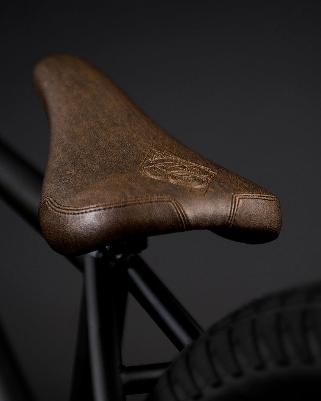 Close-up of a brown seat, engraved with the Fly Bikes logo, mounted on the sleek CrMo frame of the Neutron 20 Inch BMX bike, set against a dark background.