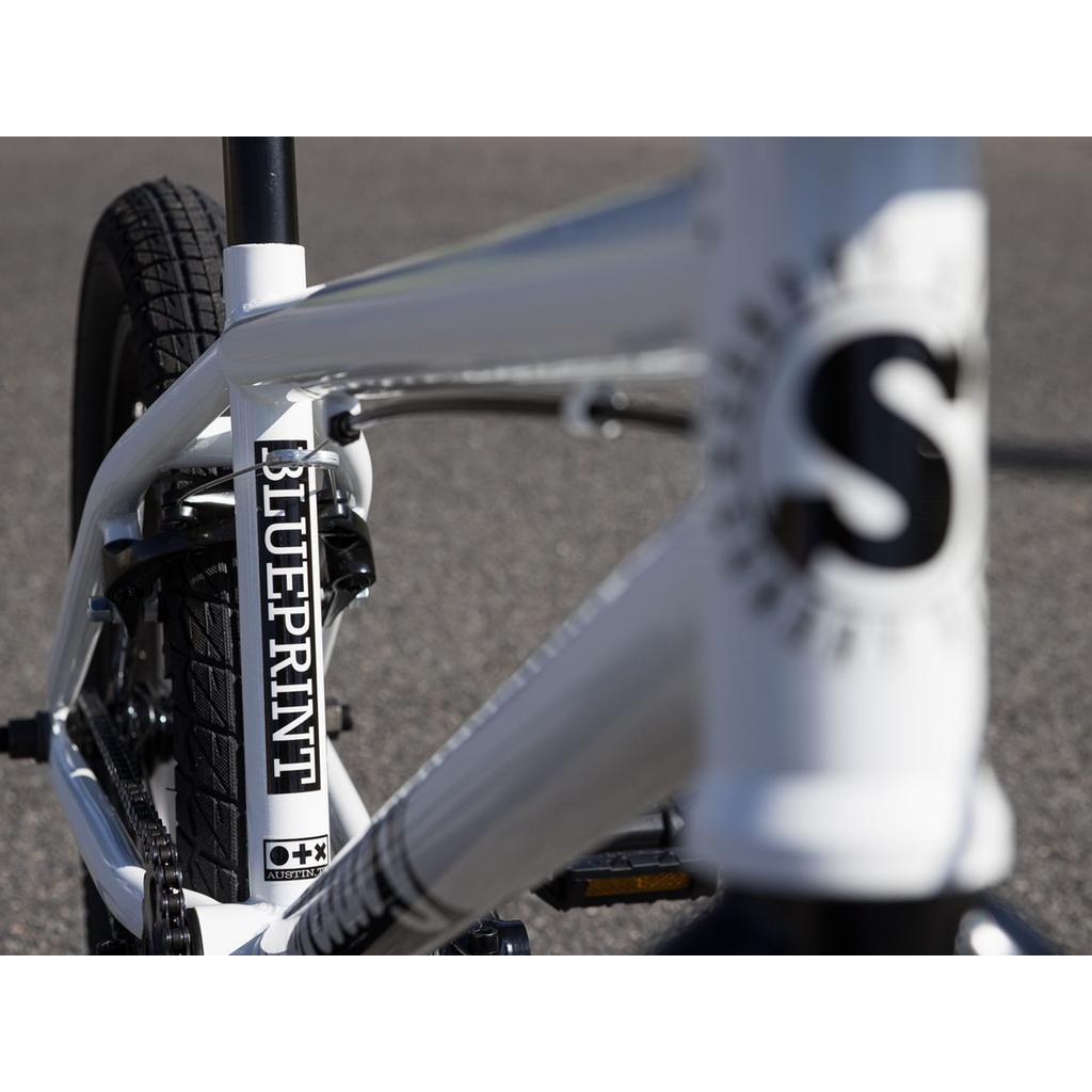 Close-up of the Sunday Blueprint 20 inch Bike beginner BMX frame showing "BLUEPRINT" on the front tube and a large "S" logo, with quality Odyssey parts and asphalt in the background.