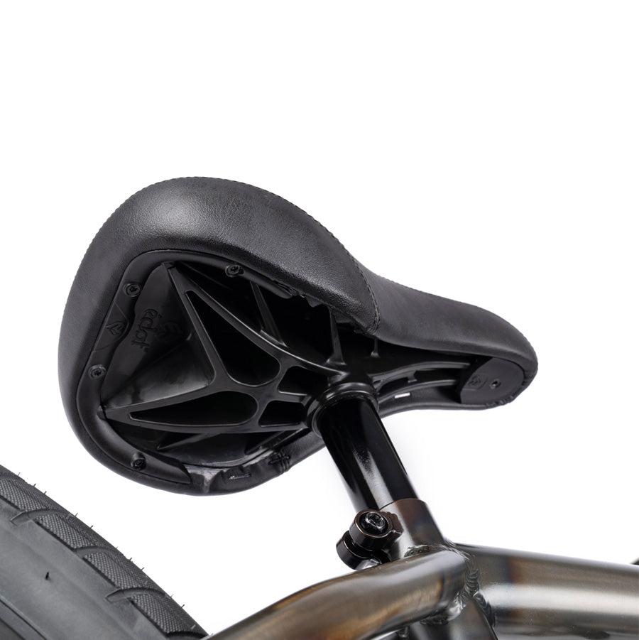 Close-up view of a black bicycle seat with a triangular, supportive underside structure attached to a metallic frame, highlighting part of the rear wheel. This Wethepeople Justice 20 Inch Bike features top-of-the-line BMX components that ensure durability and performance.