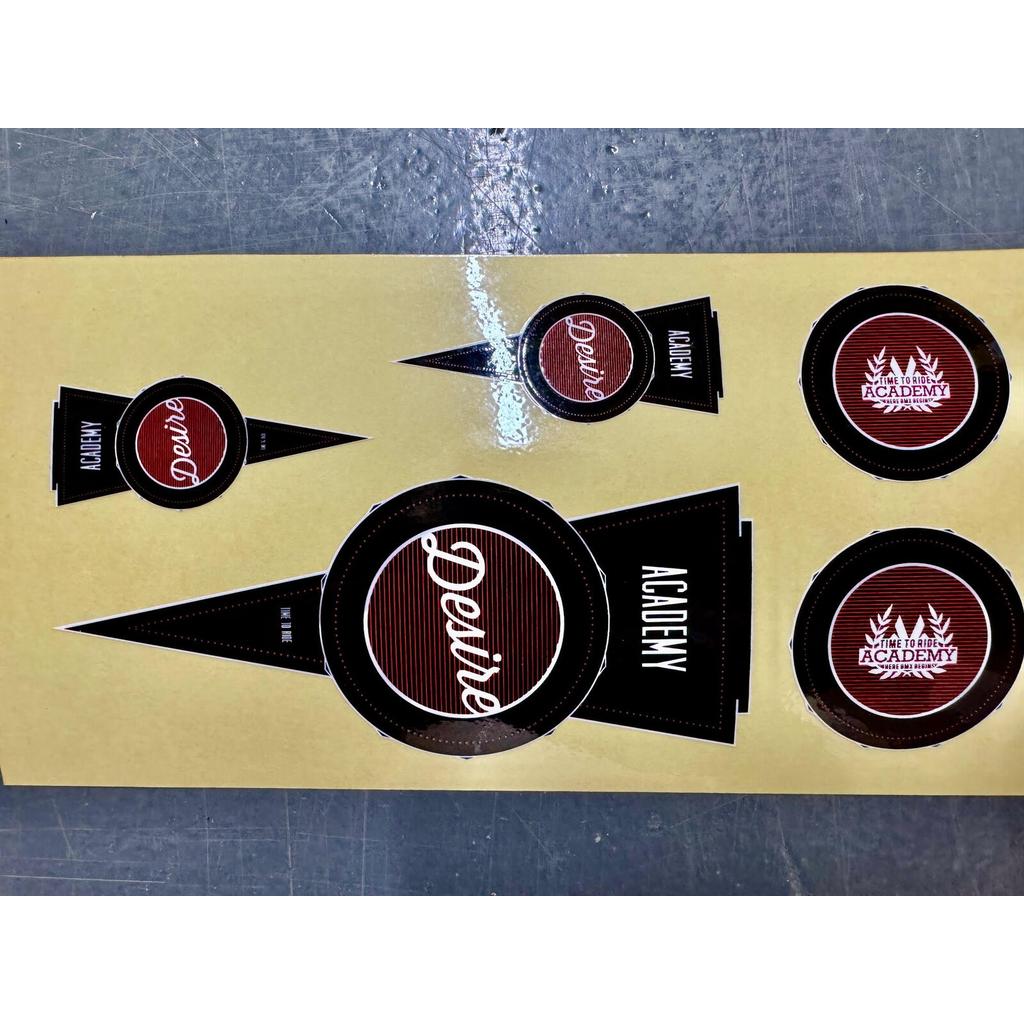 Academy Desire Sticker Pack featuring a sheet of five replacement stickers with red and black circular designs, including the text “Desire,” “Academy,” and “Time to ride,” for 2016 Purple / White models.