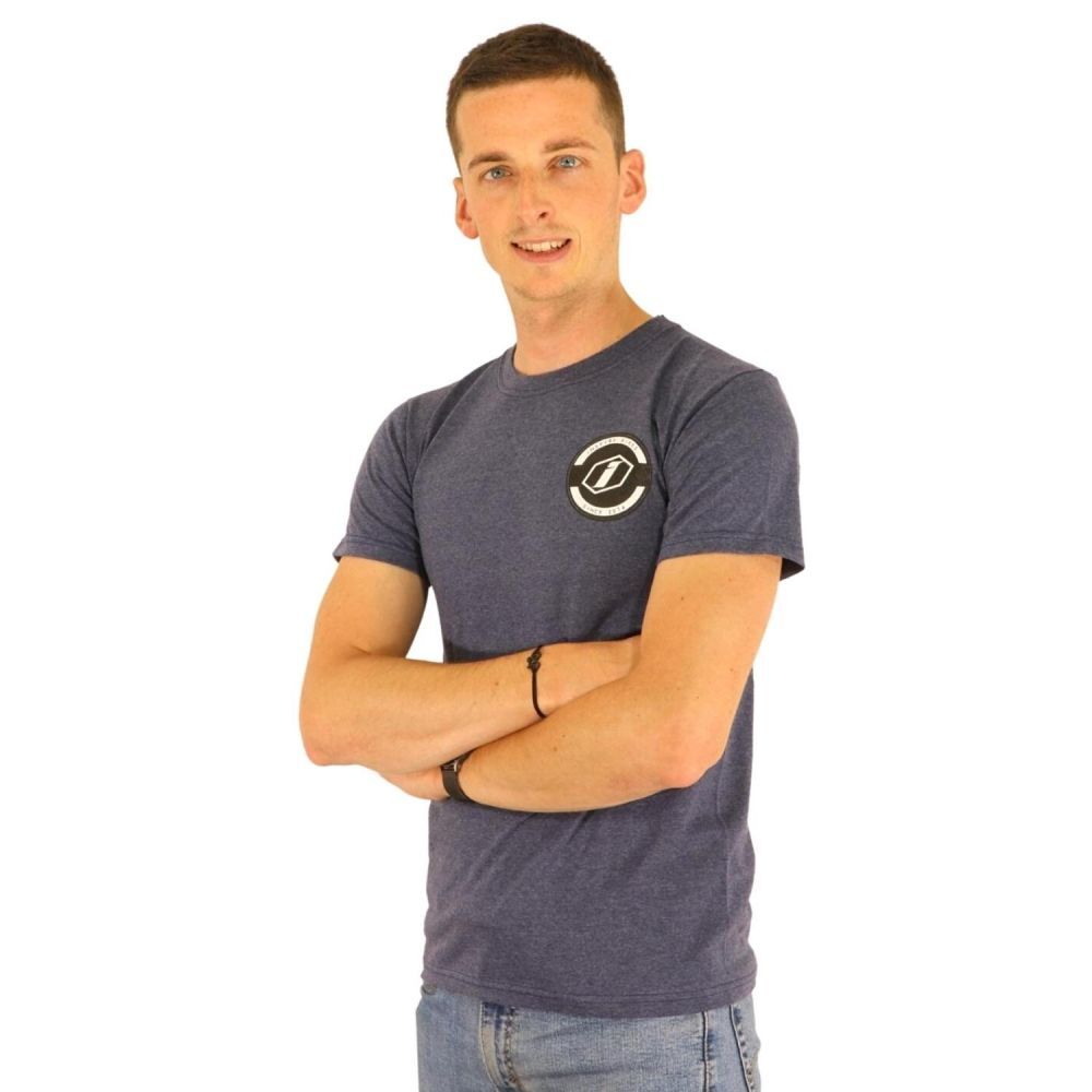 A person stands with arms crossed, smiling at the camera, wearing the Inspyre Icon T-Shirt in navy blue 100% cotton with a small circular logo on the left chest, paired with light blue jeans.