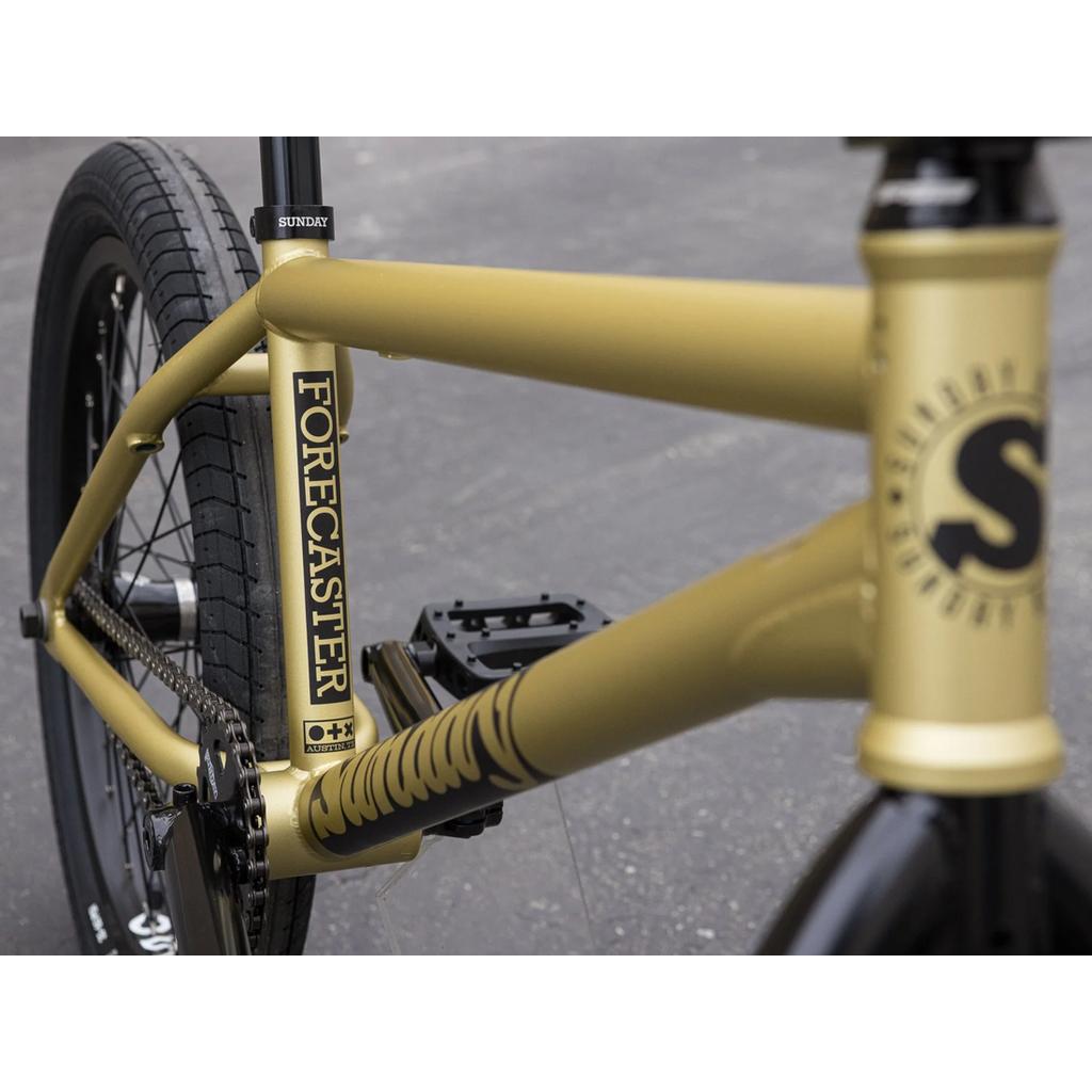 Close-up of the Sunday Forecaster 20 Inch Bike (2025) / Broc Raiford in matte gold, showing "Forecaster" and "Sunday" branding, rear wheel with Odyssey Clutch v2 Freecoaster, pedals, and part of the handlebar.