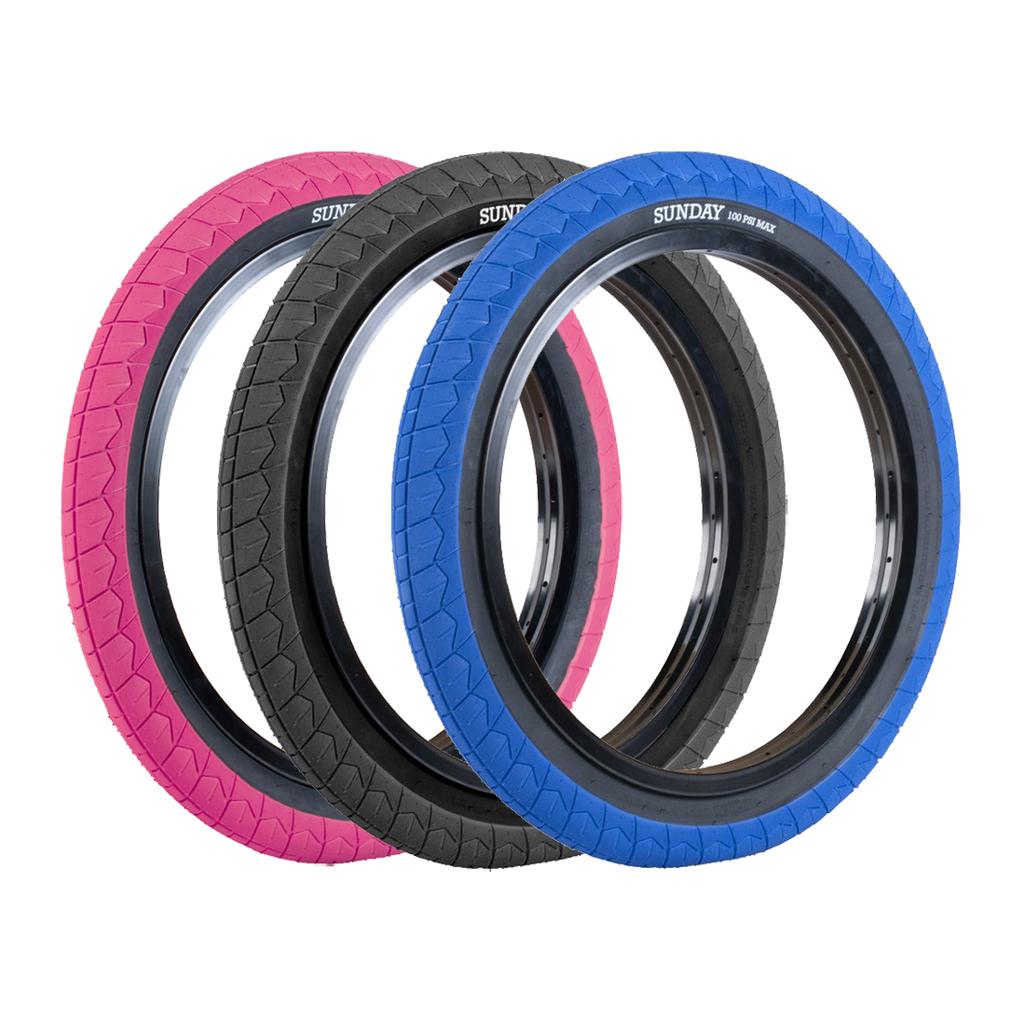 Three Sunday Current V2 Tyres in pink, black, and blue stand upright side by side, each showing the brand name. These affordable tyres feature a grippy tread for excellent control on every ride.