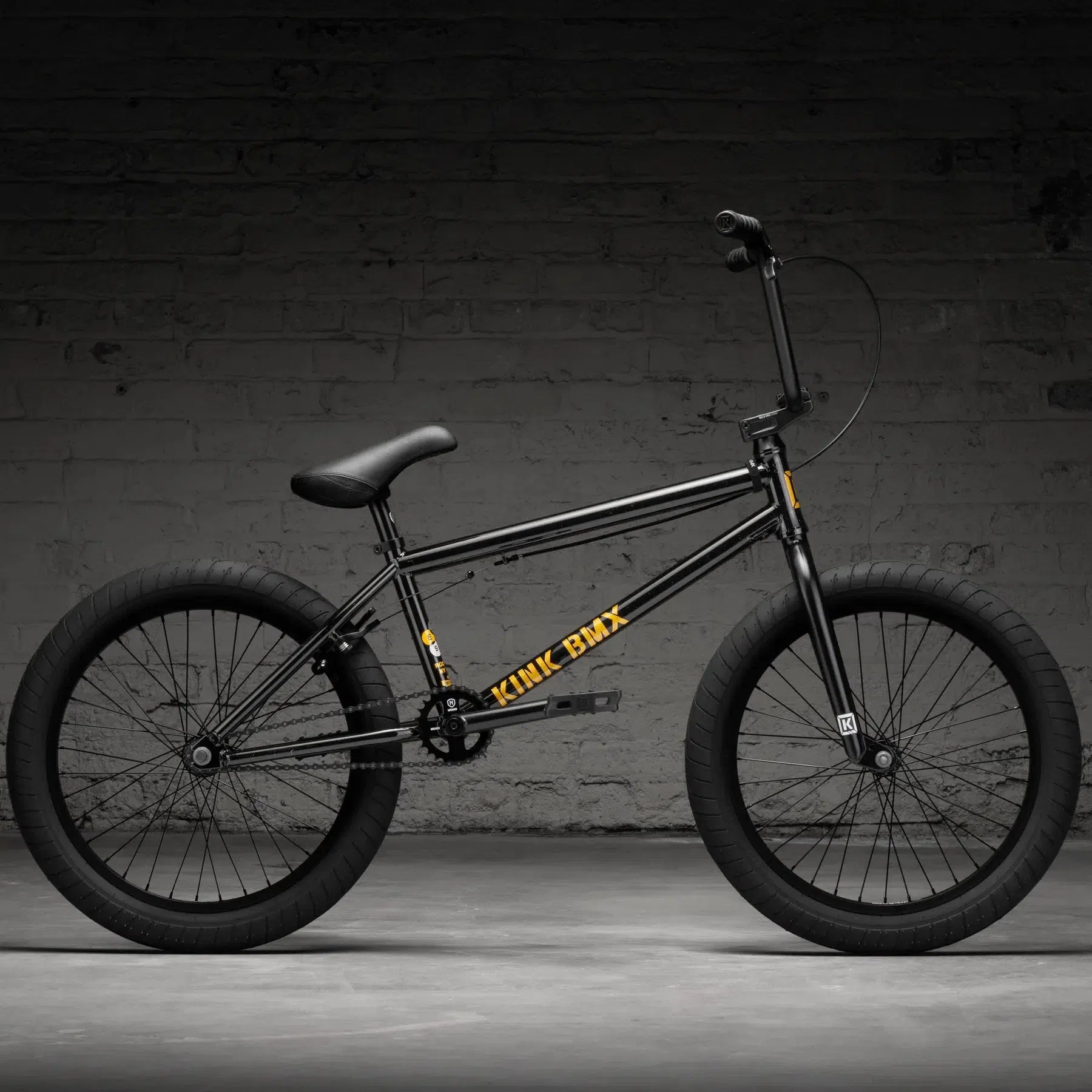 The Kink Gap 20 Inch Bike (2026), a black BMX featuring yellow lettering, made from durable chromoly steel, is set against a dark brick wall backdrop.