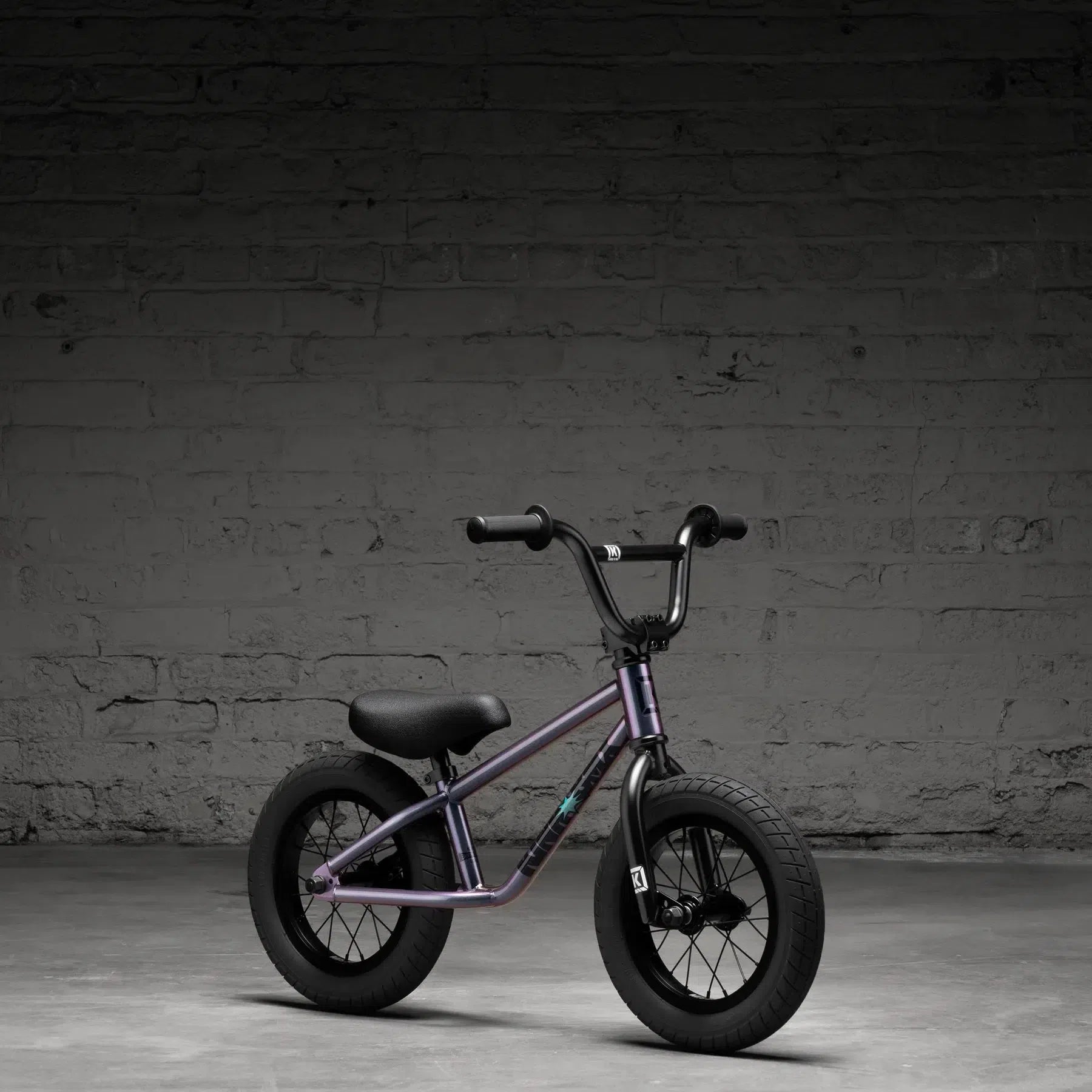 A compact purple Kink Coast 12 Inch Balance Bike (2026) with thick tires effortlessly leans against a textured gray brick wall on concrete, embodying its rugged style.
