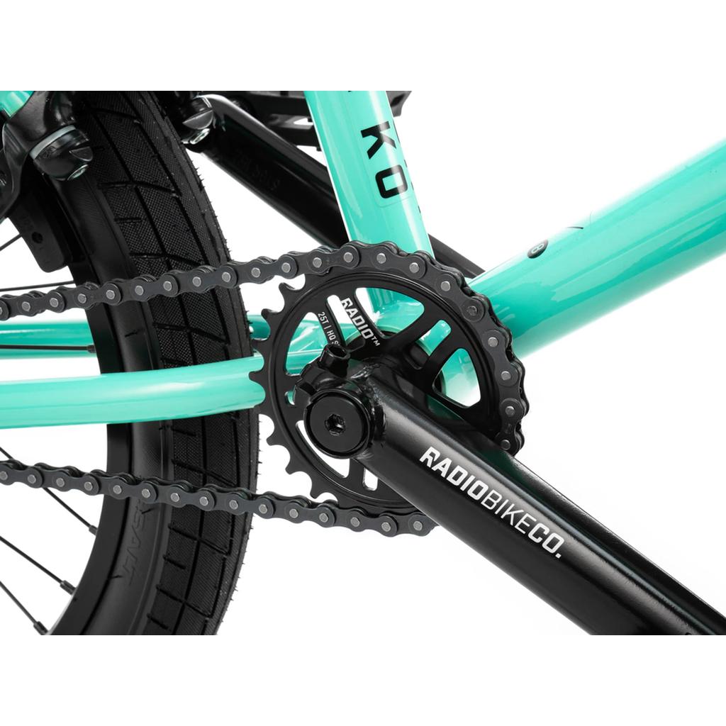 Close-up of a turquoise Radio Saiko 18 Inch Bike frame featuring a black pedal crank, chainring, chain, and rear tire with Salt Tracer Tyres. The crank arm displays the "RADIOBIKECO" logo.