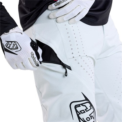 A person wearing the TLD 25.1 Sprint Pant (Youth) in Chalk, crafted from durable materials, unzips the pocket. Their white gloves and black-accented attire complement their streamlined look, prepping them for action.