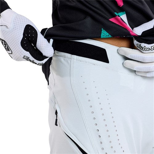 A person in a black and pink jersey with white gloves adjusts their TLD 25.1 Sprint Pant (Youth) in Chalk, featuring durable perforations and a handy side zipper—ideal for any daring sprint kit.