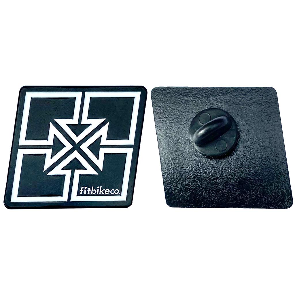 The Fit Bike Co Key Enamel Pin is shown: one side displays a white geometric cross with "fitbikeco." text, while the other side shows the black metal back and clasp.
