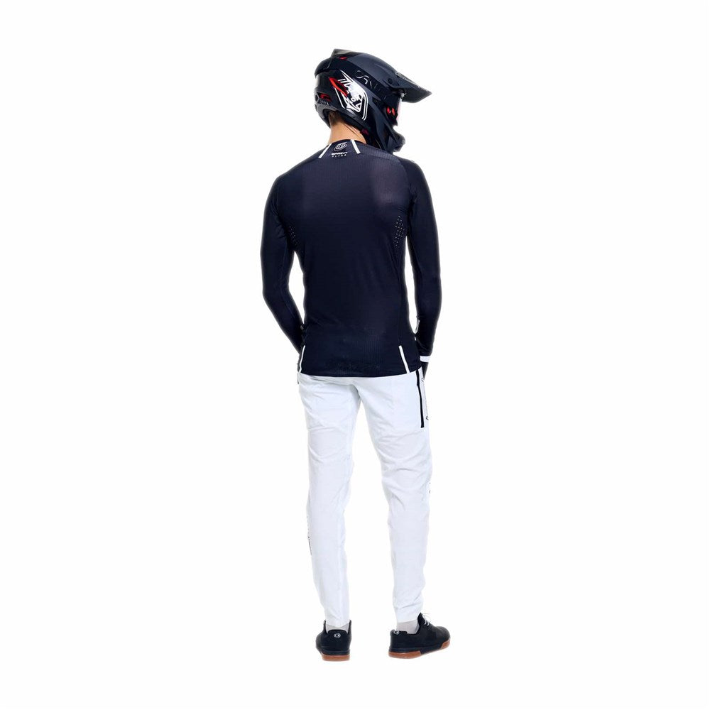 A person stands facing away from the camera against a plain white background, wearing a black helmet, black long-sleeved shirt, and TLD 26.1 Sprint Ultra Pant / Mono White with a lightweight design.
