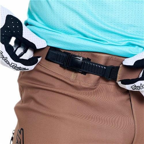 Close-up of a person wearing the TLD 26.1 Sprint Ultra Pant / Mono Sienna, a light blue shirt, and black-and-white gloves, adjusting a black belt buckle at the waist to showcase Troy Lee racing gear’s lightweight design.