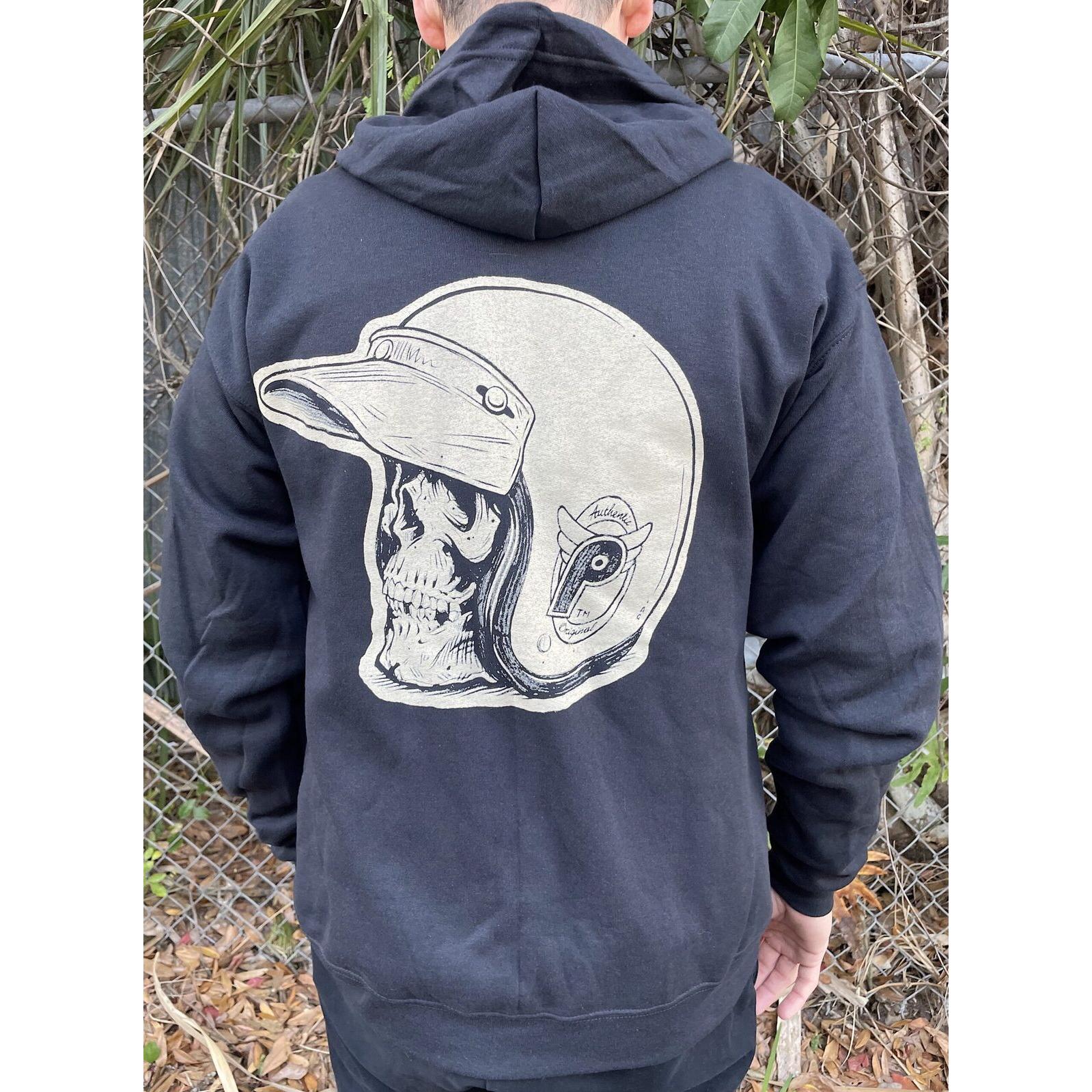 Wearing the Profile From the Dungeon Zip Hoodie, featuring a large skull in a motorcycle helmet by Adi Gilbert on the back, this person stands before a chain-link fence and foliage.