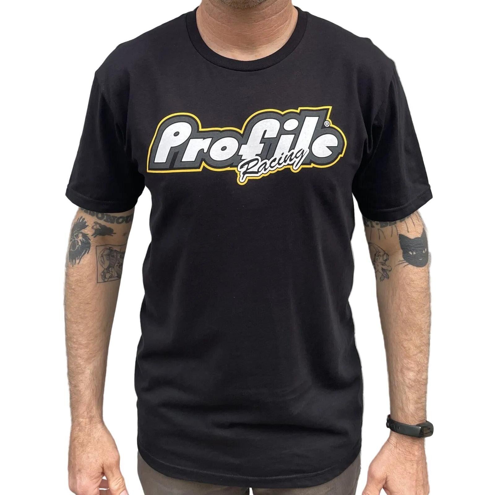 A person in a black Profile F1 Short Sleeve T-Shirt, featuring F1 Hubs, stands before a gray metal wall with tattoos visible on both arms.