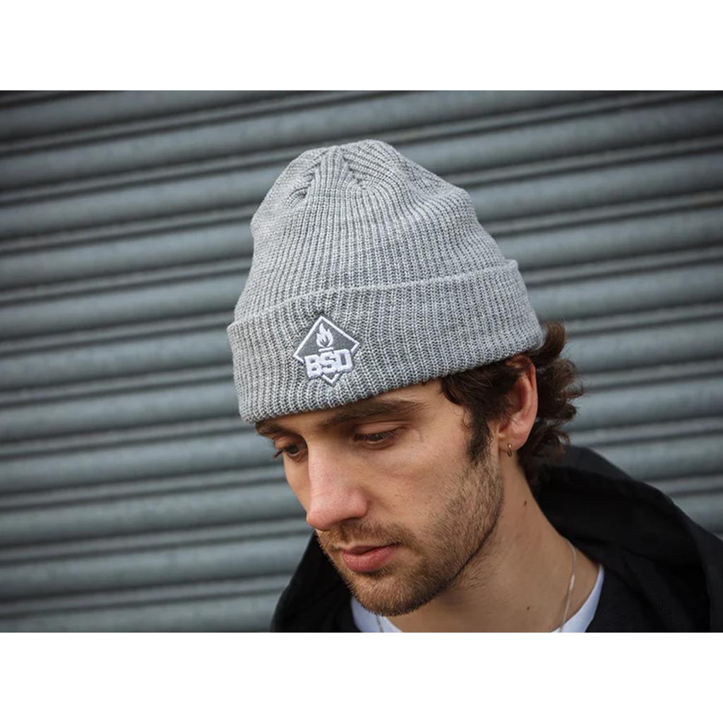 A person in a BSD Roastin Beanie and a black jacket stands before a corrugated metal backdrop.