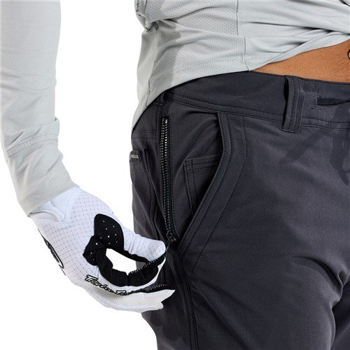 Wearing TLD 25.2 Ruckus Long Travel Pants in Mono Carbon, a person in a long-sleeve shirt one-handedly unzips a thigh pocket while wearing a white glove.