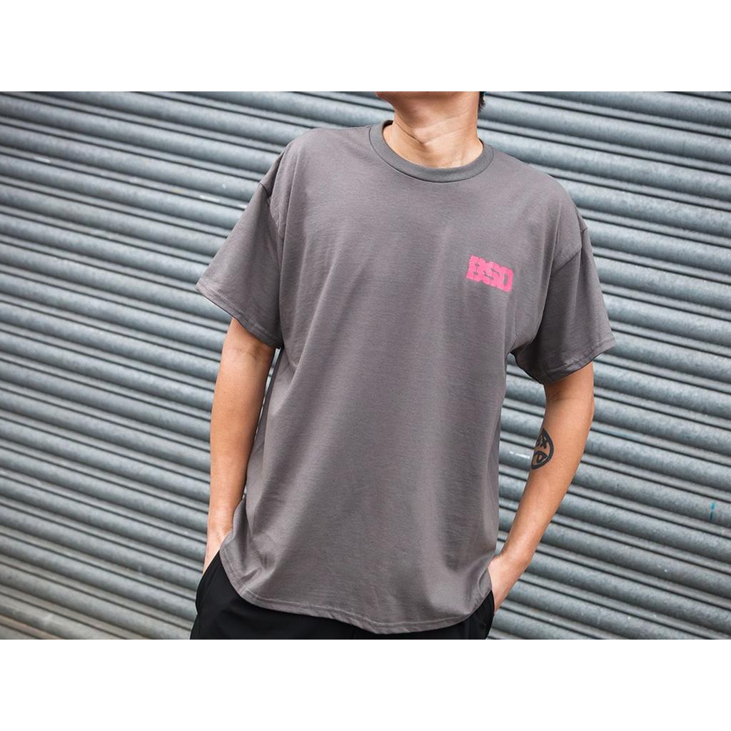 Wearing the BSD Tuned Out T-Shirt, a grey heavyweight cotton tee featuring pink "BSD" text on the chest, this person stands in front of a metal roller shutter with hands in pockets for a streetwear vibe.