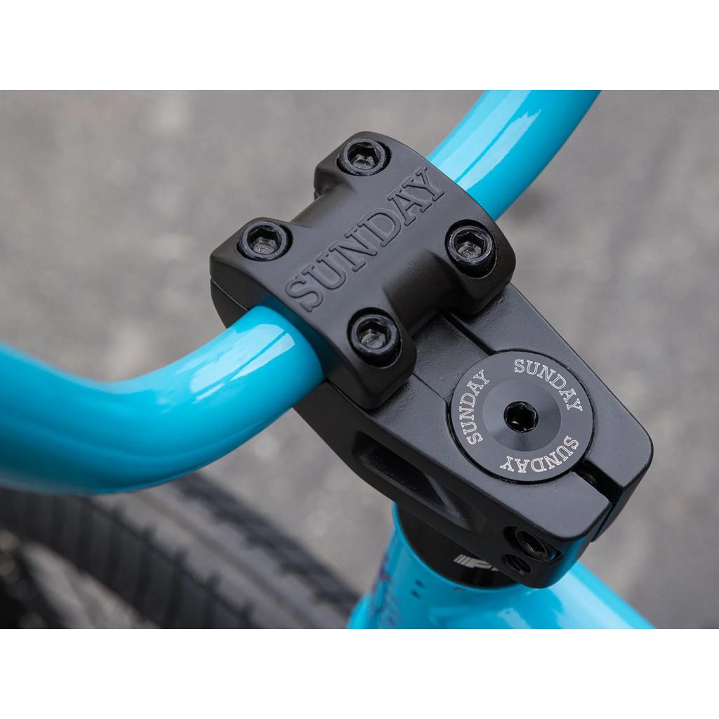 Close-up of a black handlebar stem engraved with "SUNDAY," securing bright blue bars on the Sunday Forecaster 20 Inch Bike (2025) / Aaron Ross. Blurred pavement in the background.