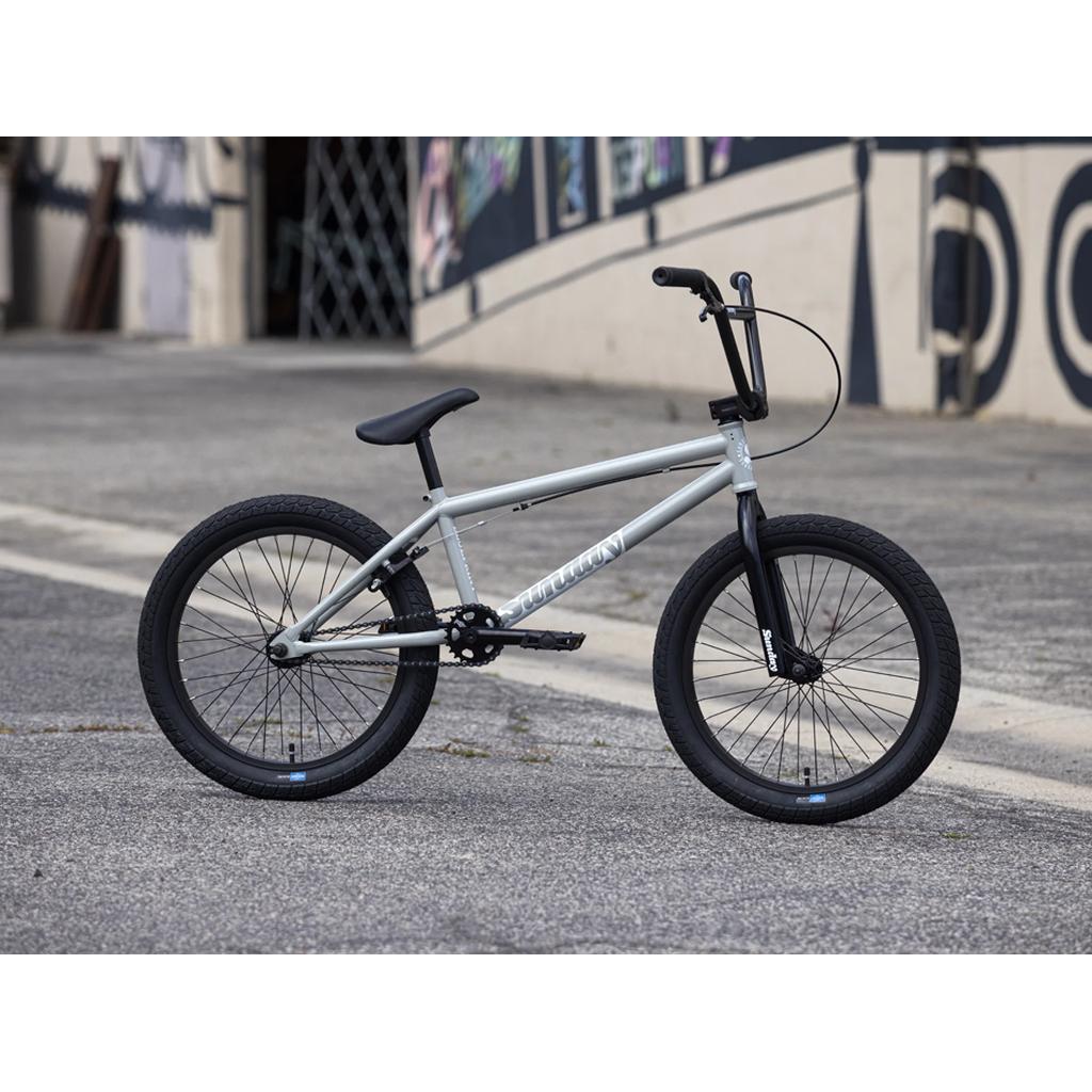A silver Sunday Blueprint 20 inch Bike with black tires and handlebars rests on a paved surface in front of a wall displaying abstract art. Designed with pro-level geometry, the Sunday Blueprint 20 inch Bike delivers both style and performance, making it an ideal choice for beginner riders ready to elevate their BMX skills.