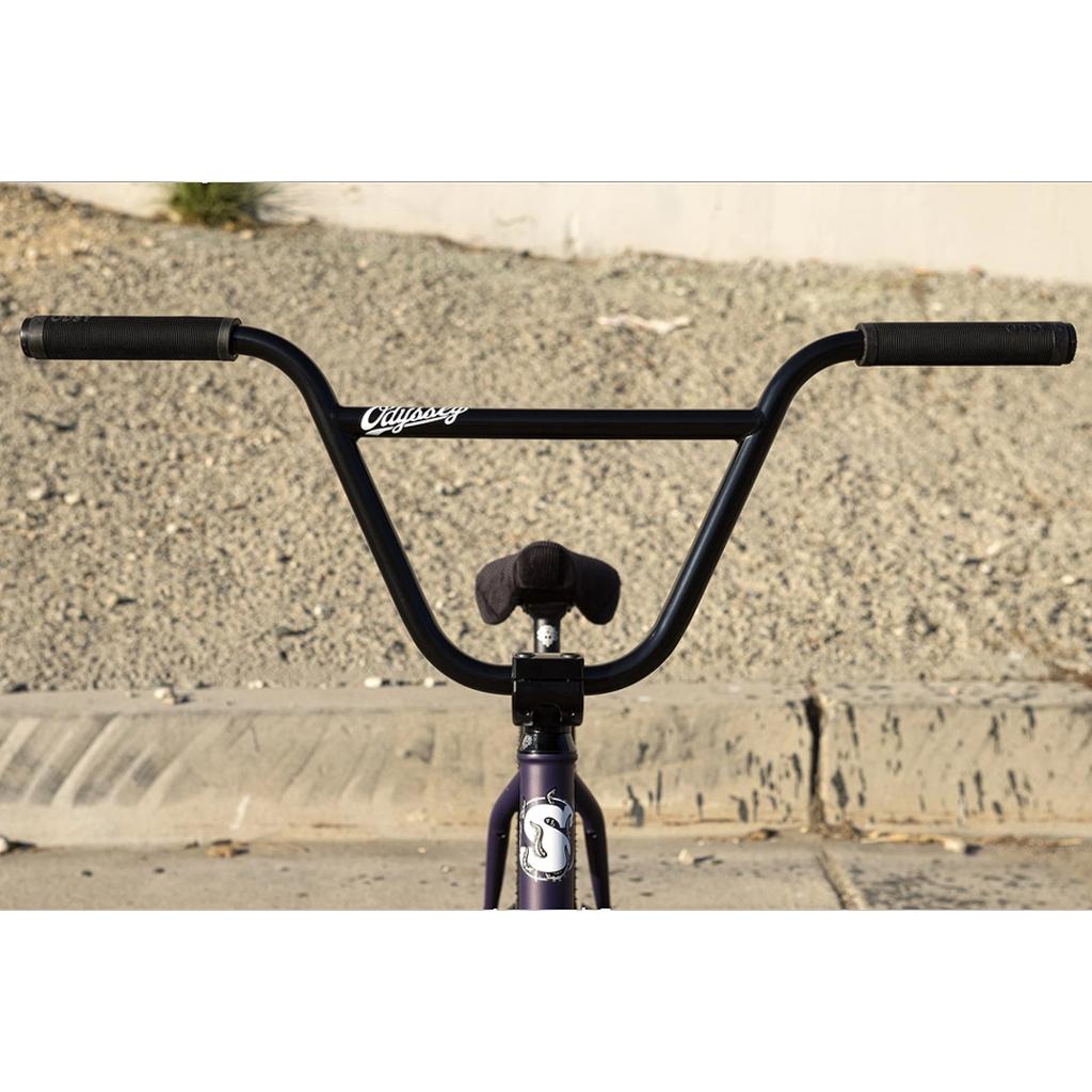 Close-up of the Sunday Dark Wave Authentic 20 Inch Bike’s black handlebars and seat, photographed from the front with a cement curb and gravel behind. This model features a 100% Chromoly frame for outstanding strength.