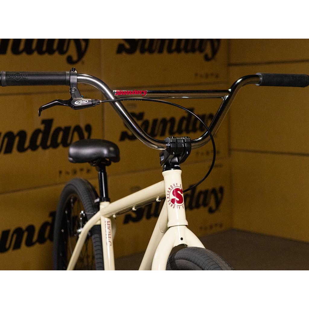 Close-up of the handlebars, stem, and front frame of the Sunday High C 29 Inch Bike, highlighting its durable chromoly construction and featuring the prominent "Sunday" logo. The background is filled with repeated "Sunday" text, emphasizing the bold style of this 29-inch cruiser.