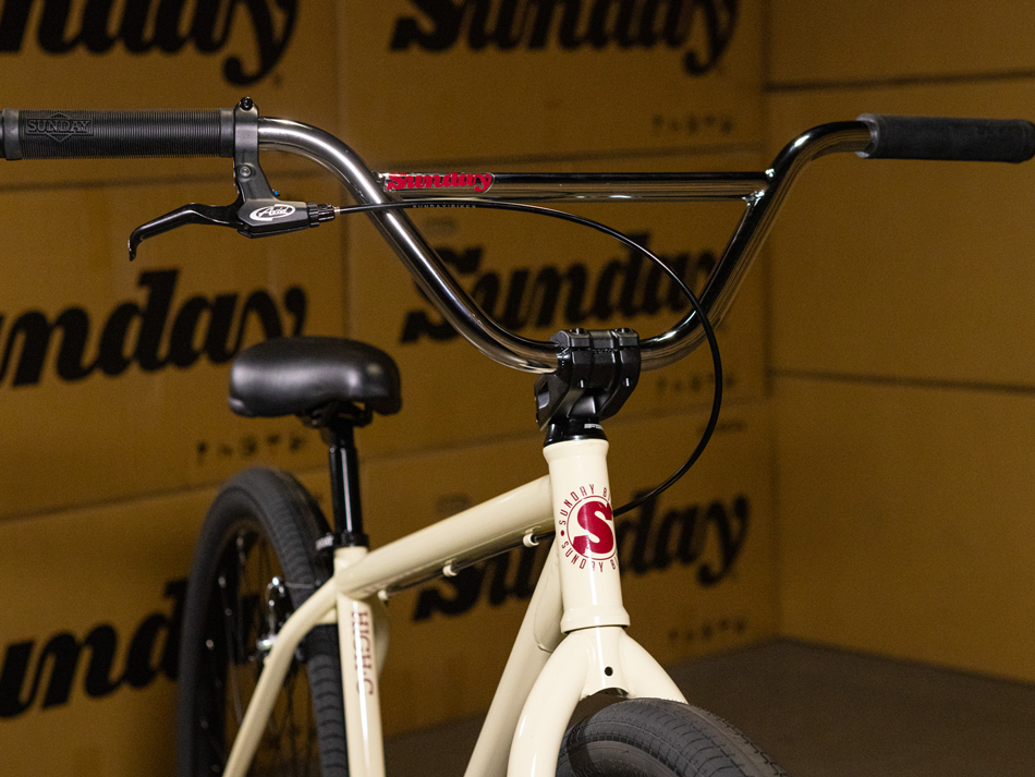 Close-up of the handlebars, stem, and front frame of the Sunday High C 29 Inch Bike, highlighting its durable chromoly construction and featuring the prominent "Sunday" logo. The background is filled with repeated "Sunday" text, emphasizing the bold style of this 29-inch cruiser.