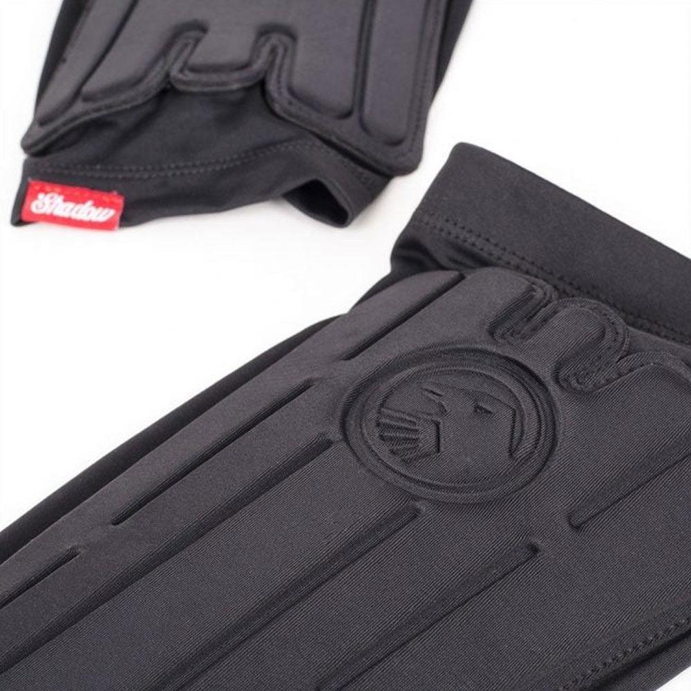 Close-up of Shadow Conspiracy Invisa-Lite Shin Pads (Pair) in black, showcasing padded construction with stitched lines, lion logo, and a visible red and white "Shadow" tag.