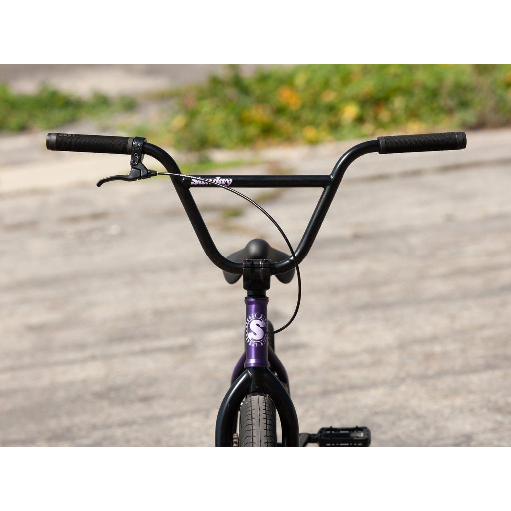 Close-up of the Sunday Scout 20 Inch Bike (2022)'s handlebars and front wheel, showing sealed hubs, a single brake lever, and a purple head tube. The bike is on a paved surface with greenery in the background.