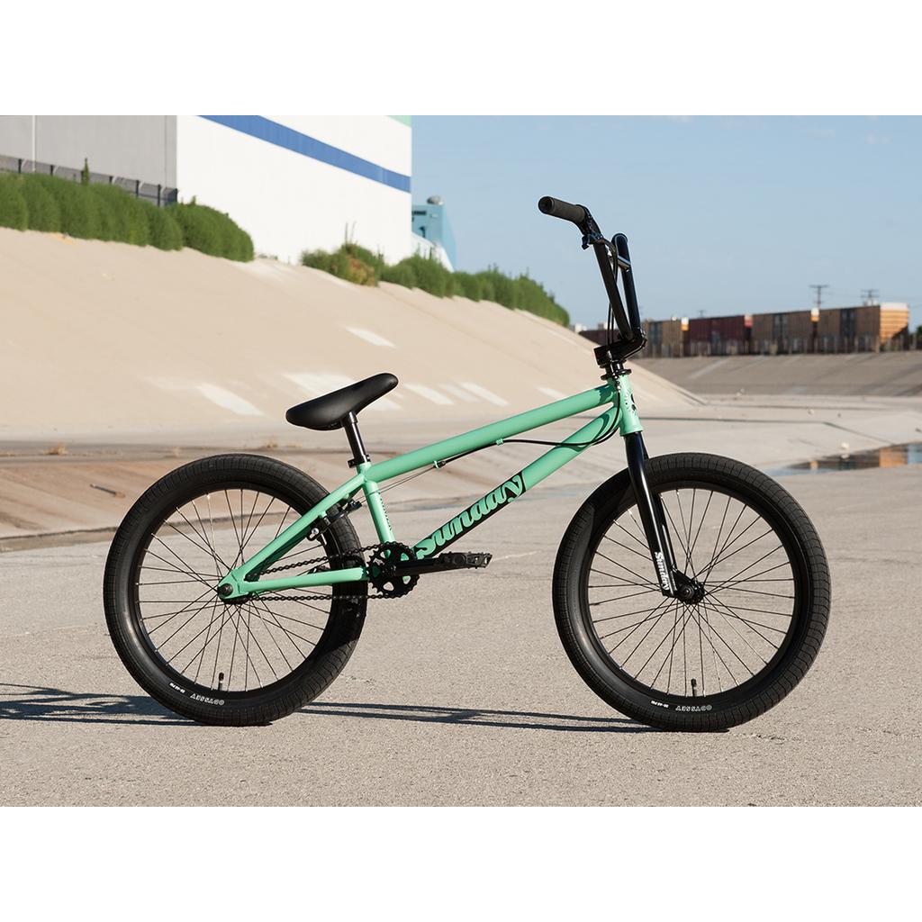 A mint green Sunday Primer Park 20 Inch Bike with black handlebars, seat, and tires is parked on pavement near a concrete embankment and several train cars.
