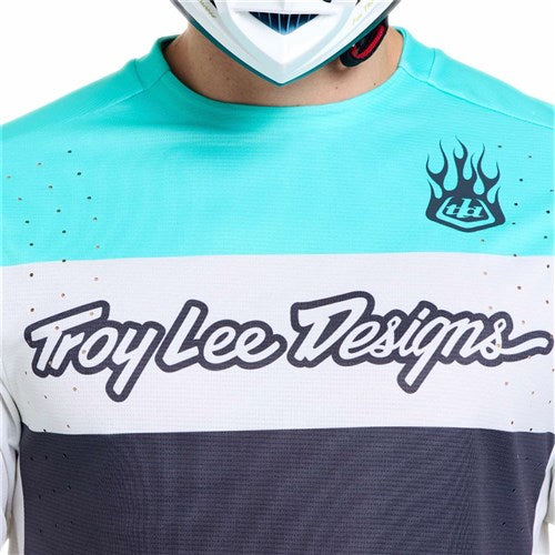 Someone wears a TLD 26.1 Sprint Pro Jersey / Yo Moto Carbon in teal, white, and gray with a Race Fit, flame logo, and chest text. Made from Bluesign approved fabrics for sustainable style by Troy Lee Designs.