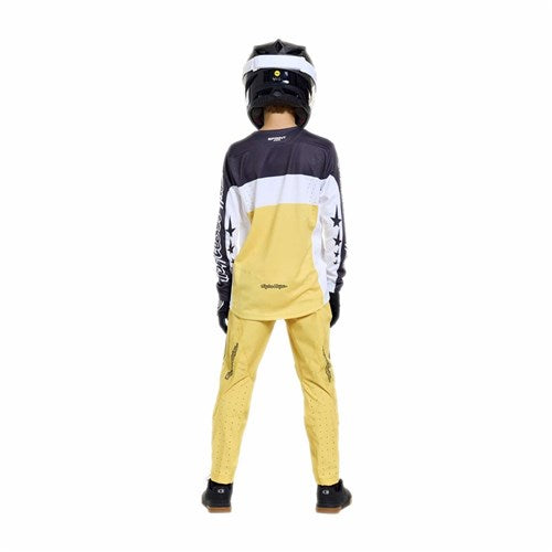 A person in a black helmet and TLD 26.1 Sprint Pro Jersey (Youth) / Yo Moto Butter stands with their back to the camera, wearing yellow riding pants, set against a plain white background.