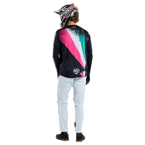 A rider in vibrant Troy Lee gear, featuring the TLD 25.1 Sprint Jersey in Full Pull Black and a striking helmet, stands with their back to us, gray pants complementing their look as they prepare for race weekends.