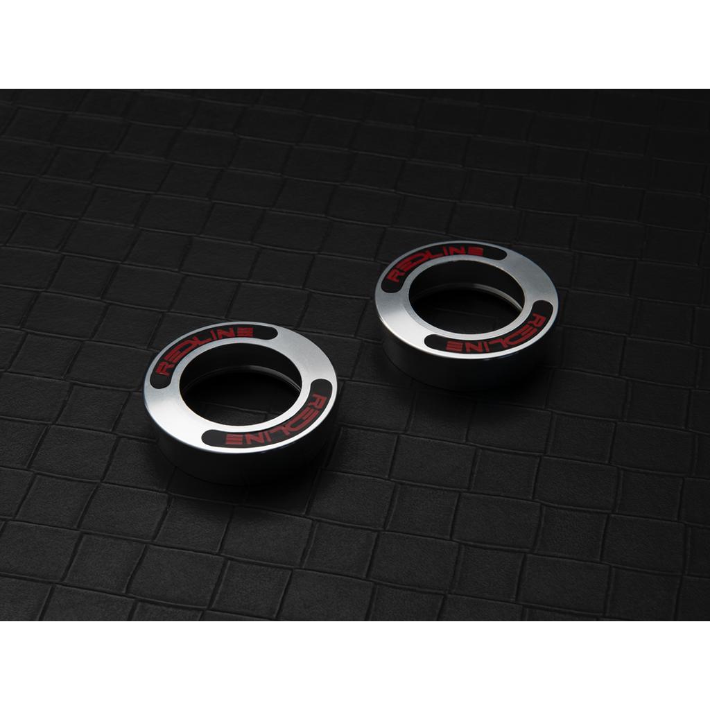 Two metallic circular objects labeled "REDLINE" rest on a textured black grid surface, resembling components like the Redline Techmatic USA BB Set.