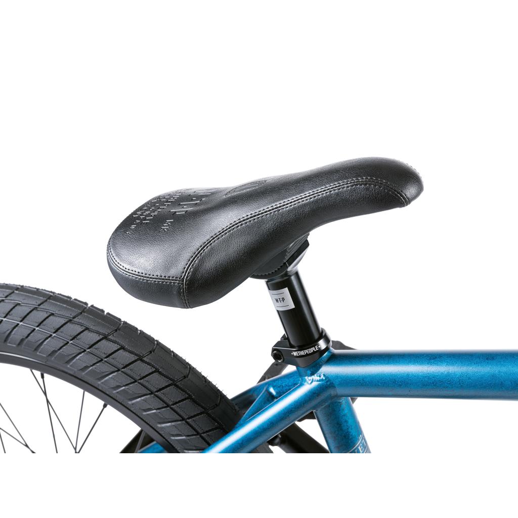 Close-up of a black bicycle seat on a blue Wethepeople Revolver 20 Inch Bike frame, partially showing the rear wheel, set against a white background.