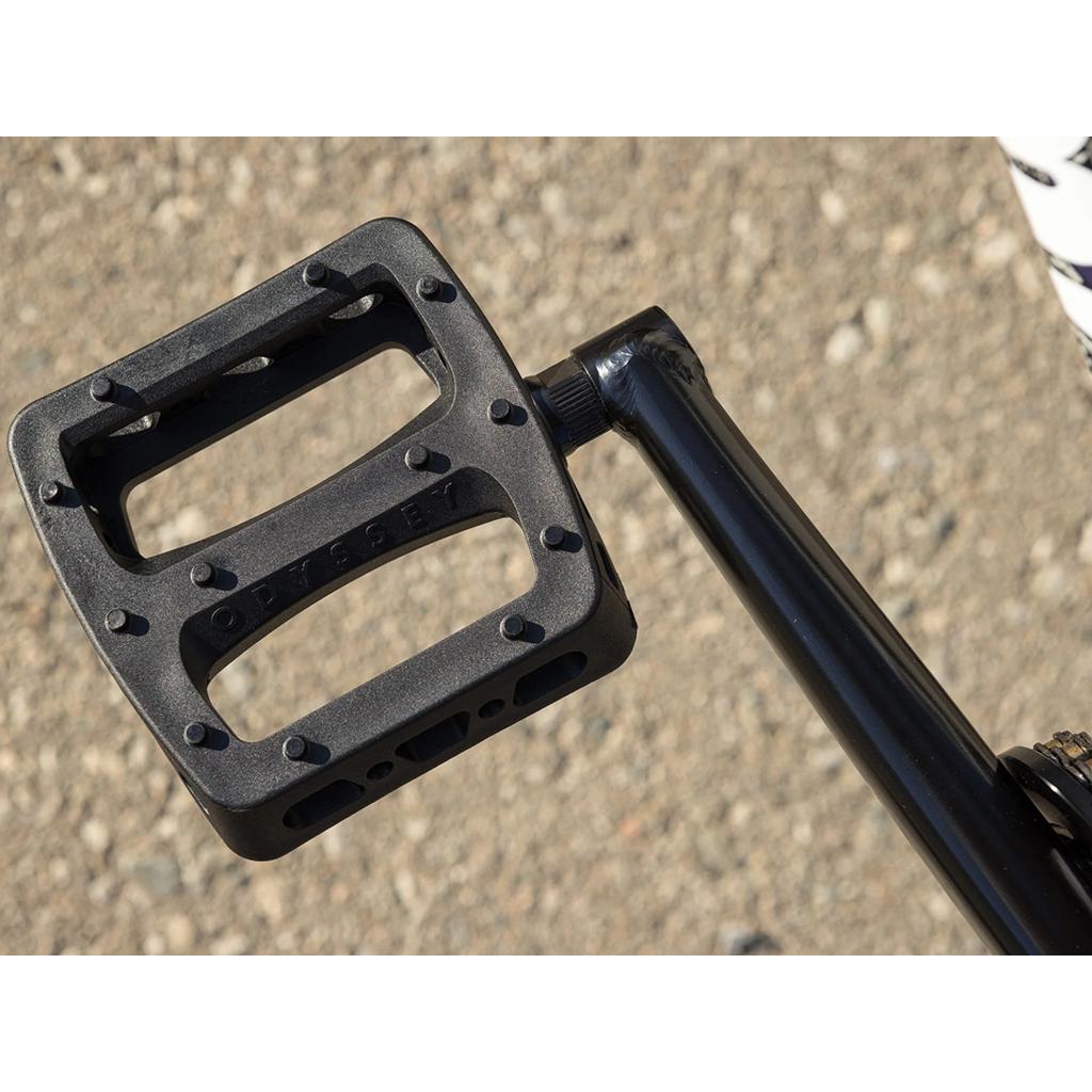 Close-up of a black Odyssey bicycle pedal attached to a crank arm on the Sunday Dark Wave Authentic 20 Inch Bike, with a textured outdoor ground surface in the background.