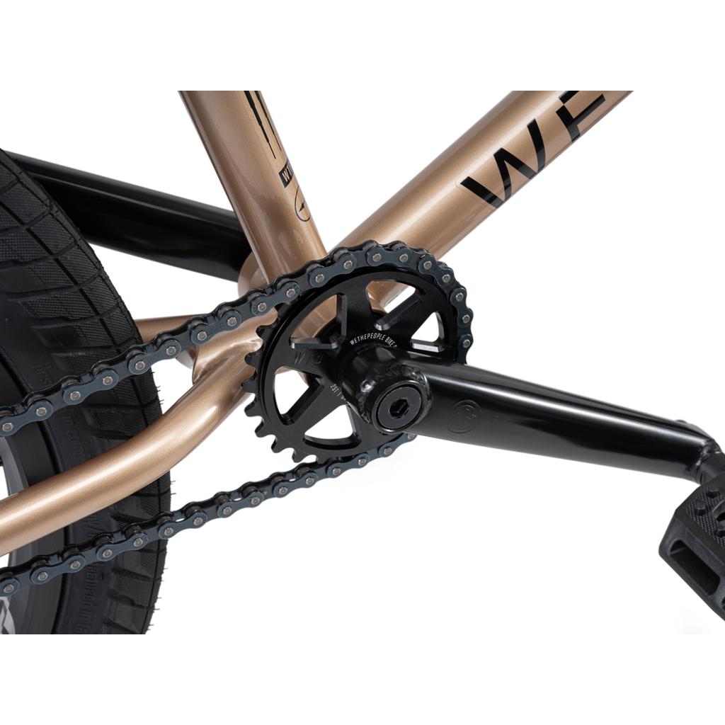 Close-up view of the crankset, chainring, chain, and pedal on a metallic gold and black Wethepeople Crysis 20 Inch BMX Bike with SaltPlus Ultra Three Piece Cranks.