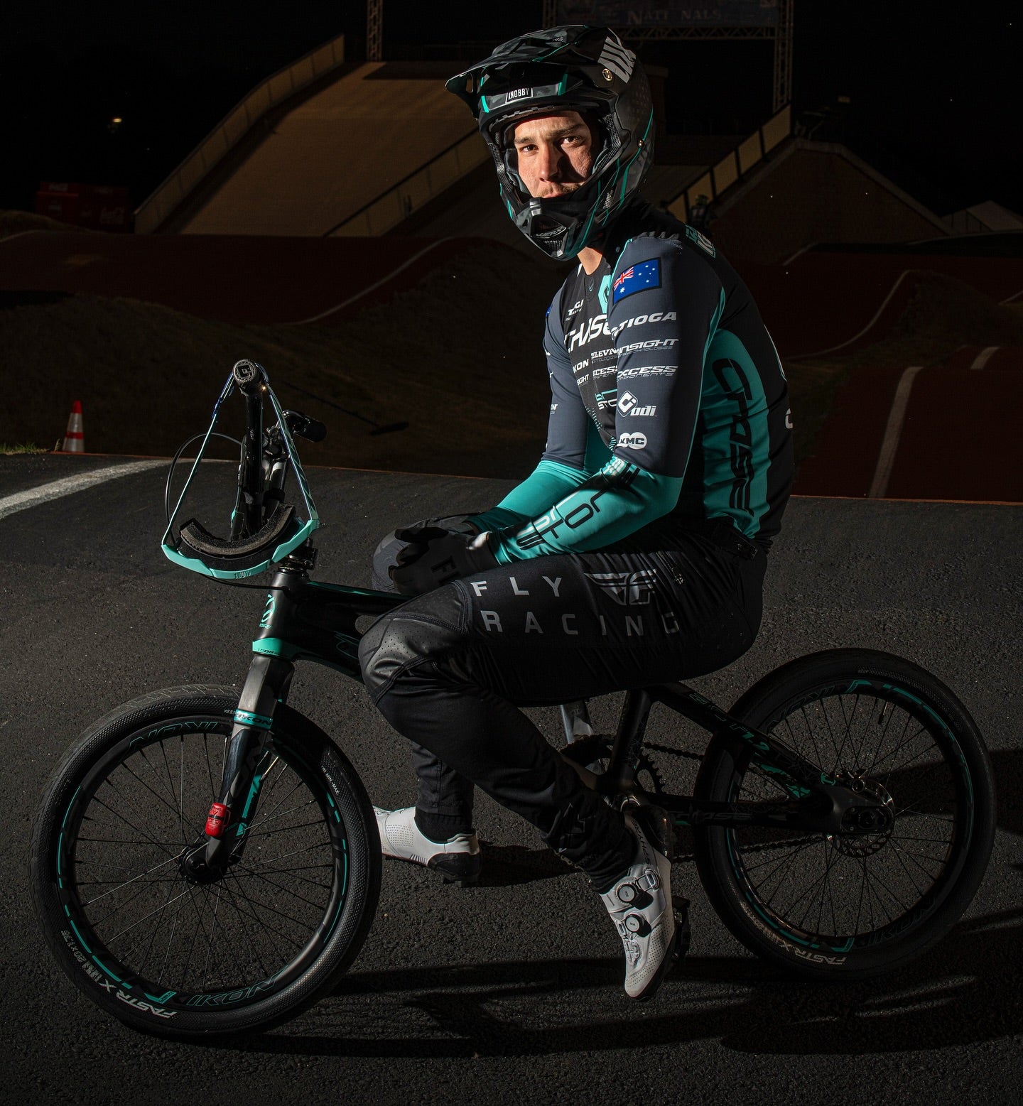 A BMX cyclist in full racing gear and a Chase Izaac Kennedy 2024 Team Replica Youth Jersey sits on a bike on an outdoor track at night, with ramps in the background, ready for BMX action.
