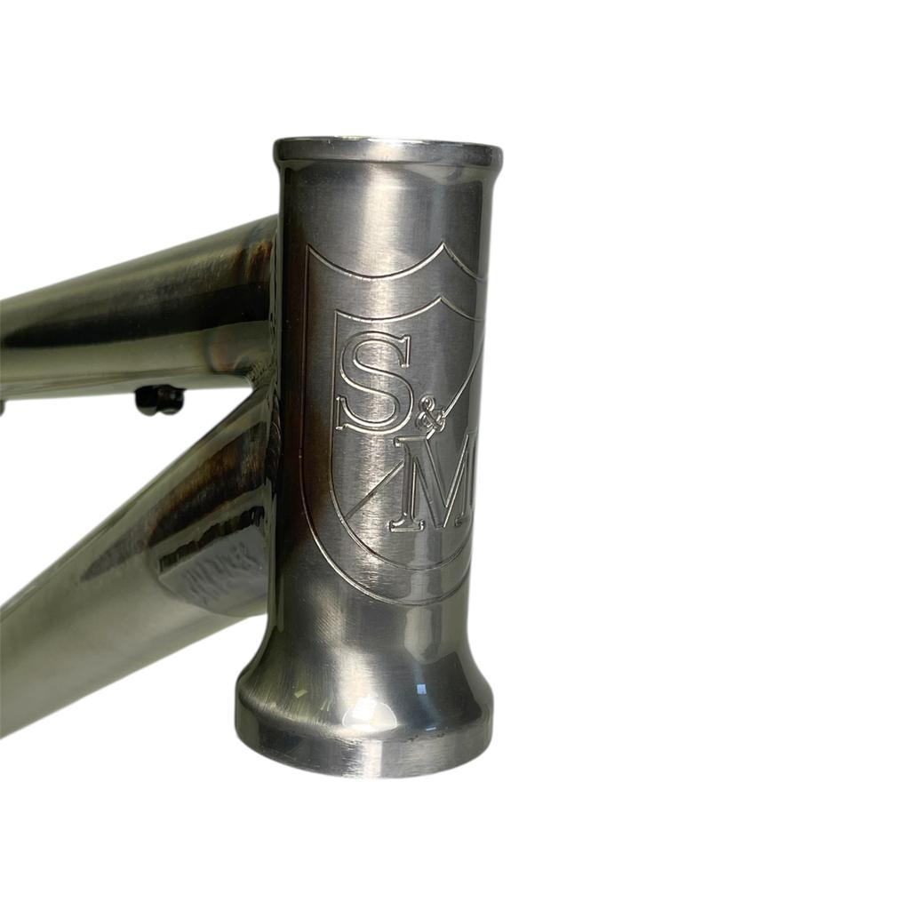 Close-up of the S&M Big Jumper V2 26 Inch Dirt Jumper Frame’s metallic head tube, showing the engraved S&M logo shield; built for disc brakes and suspension forks.