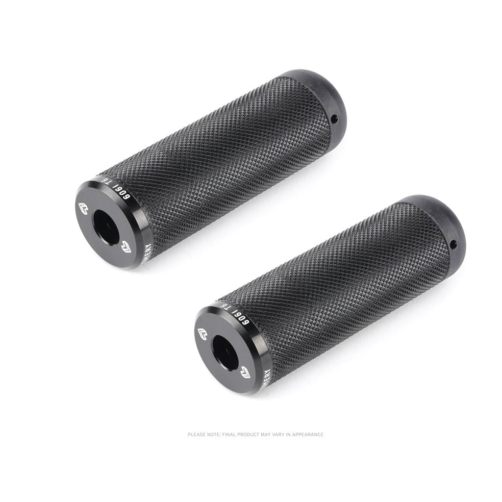 Two black, cylindrical Eclat Emery Axle Pegs with textured surfaces and metal end caps are shown on a white background, complementing alloy or flatland pegs to enhance control.