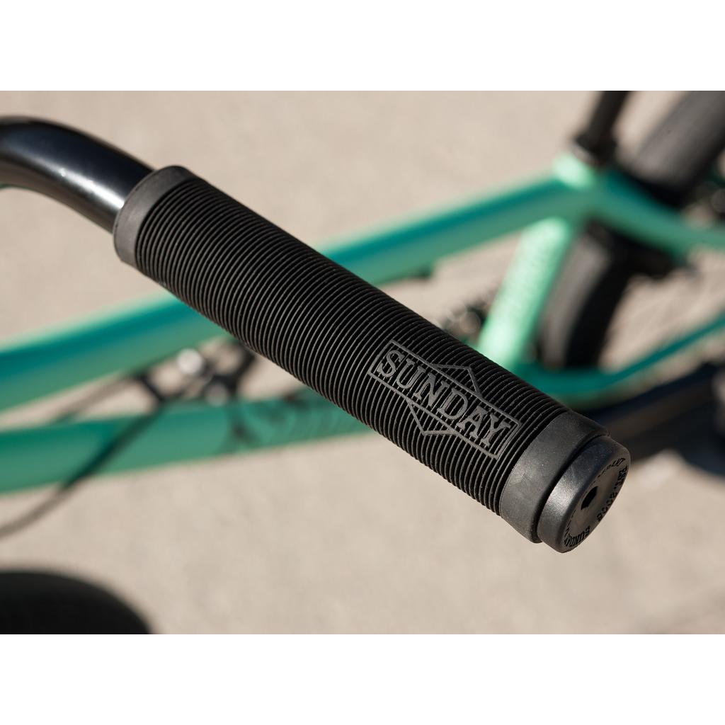 Close-up of a black ribbed handlebar grip with "SUNDAY" embossed, set against part of the teal Sunday Primer Park 20 Inch Bike frame and tire—ideal for your next park BMX session.